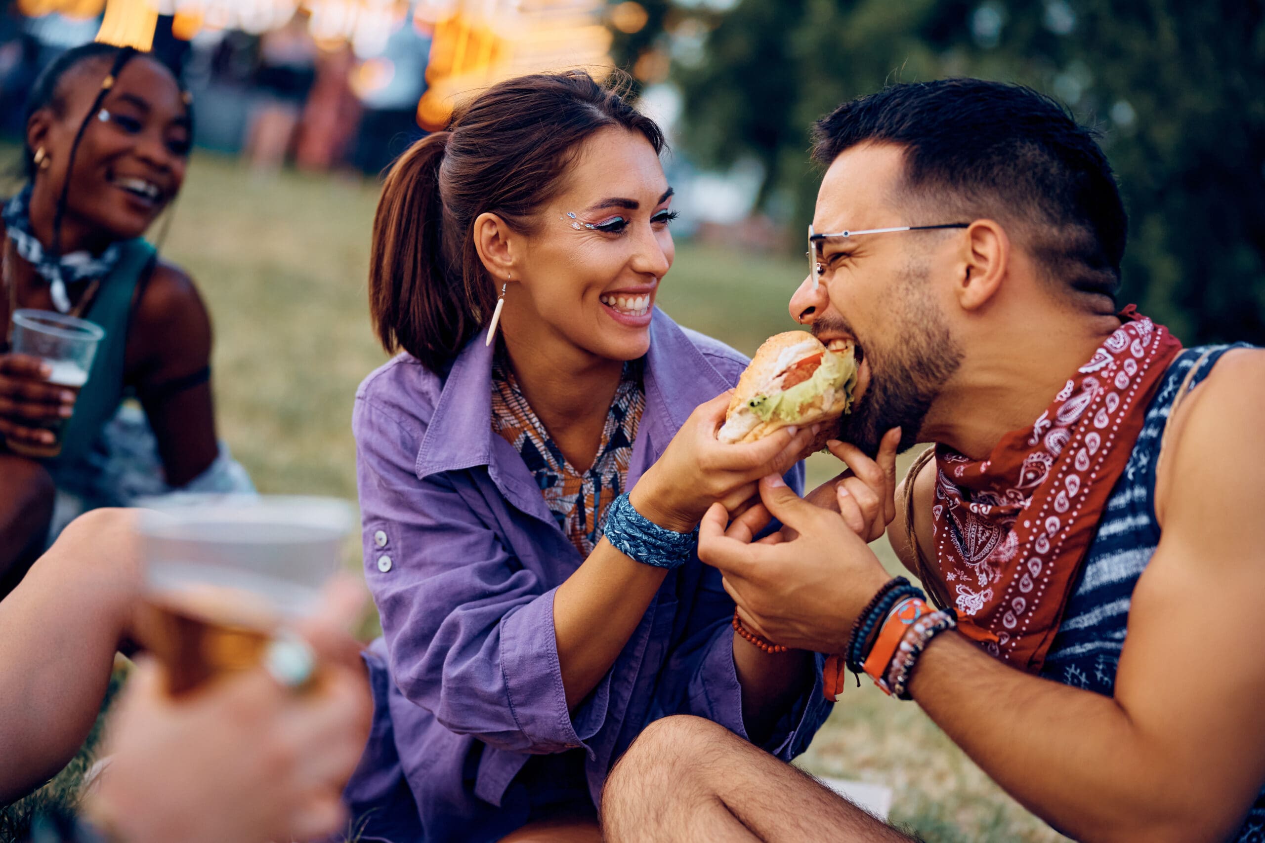 Happy couple sharing hamburger while attending summer music festival with their friends. Happy couple sharing hamburger while attending summer music festival with their friends.